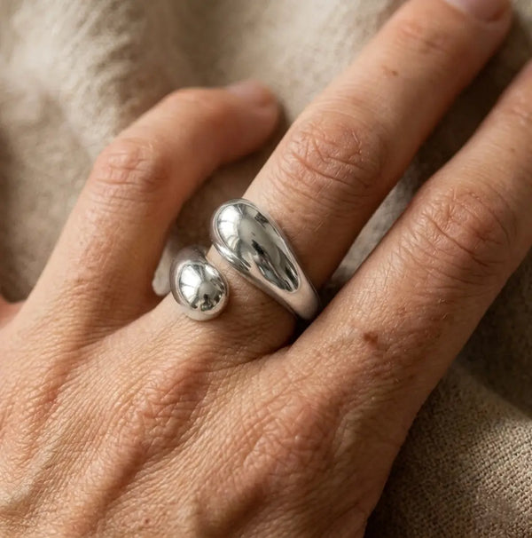 Bague argentée sculpturale Alya Audacieuse portée au doigt. Design organique en forme de goutte avec fini miroir sur fond textile beige.