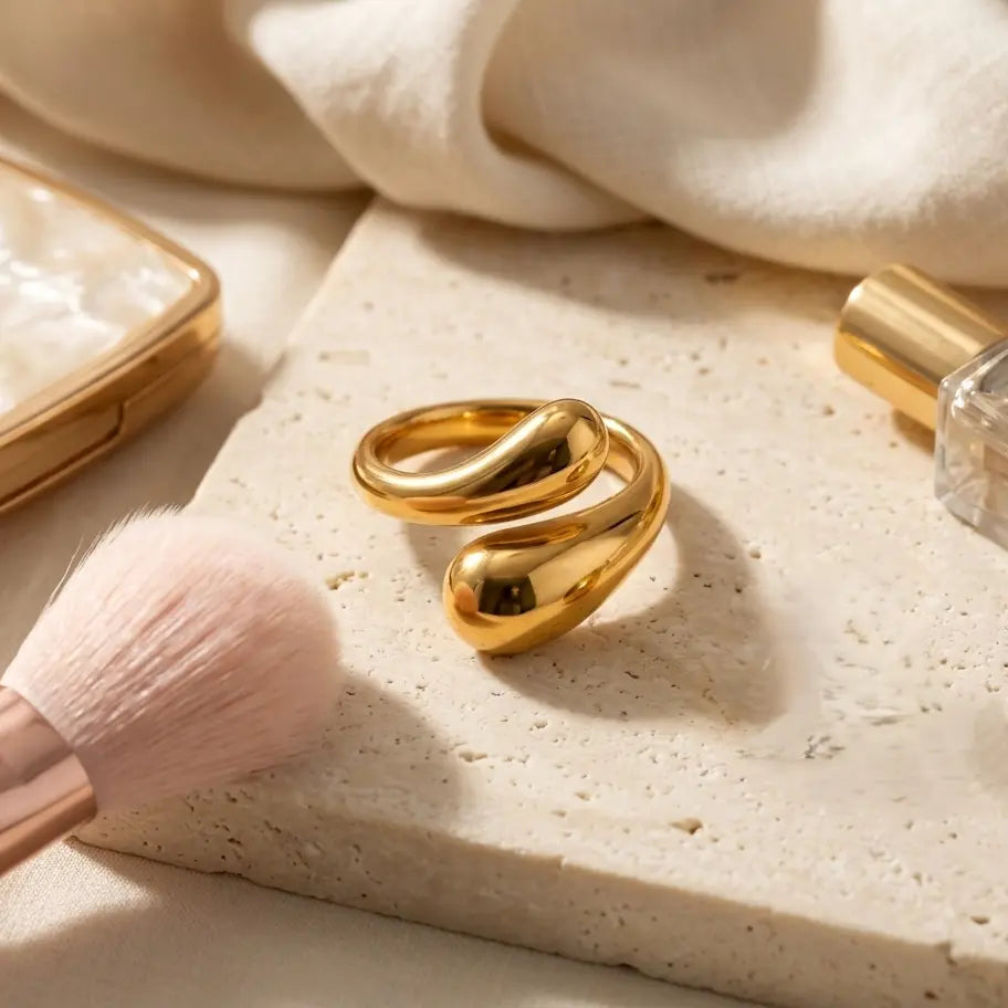 Mise en scène lifestyle de la bague dorée Alya Audacieuse sur un socle en pierre naturelle avec accessoires de beauté en arrière-plan.