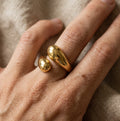 Bague dorée sculpturale Alya Audacieuse portée au doigt. Design organique en forme de goutte avec fini miroir sur fond textile beige.