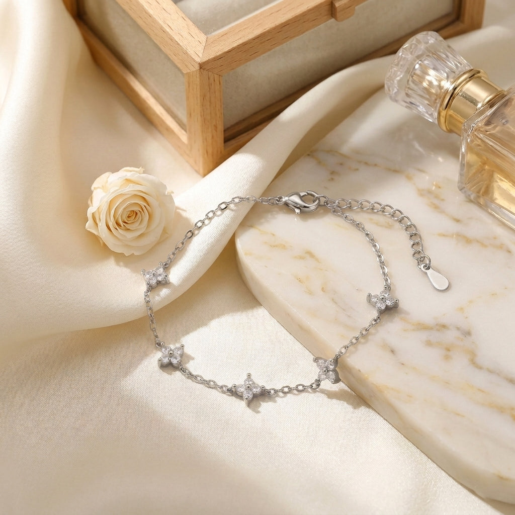 Bracelet multi-trèfles argenté Margaux Fine mis en scène dans un décor chic. Le bijou scintille sur un support minéral texturé, contrastant avec la douceur d'un tissu satiné crème et des pétales de fleurs séchées baignés dans une lumière solaire douce.