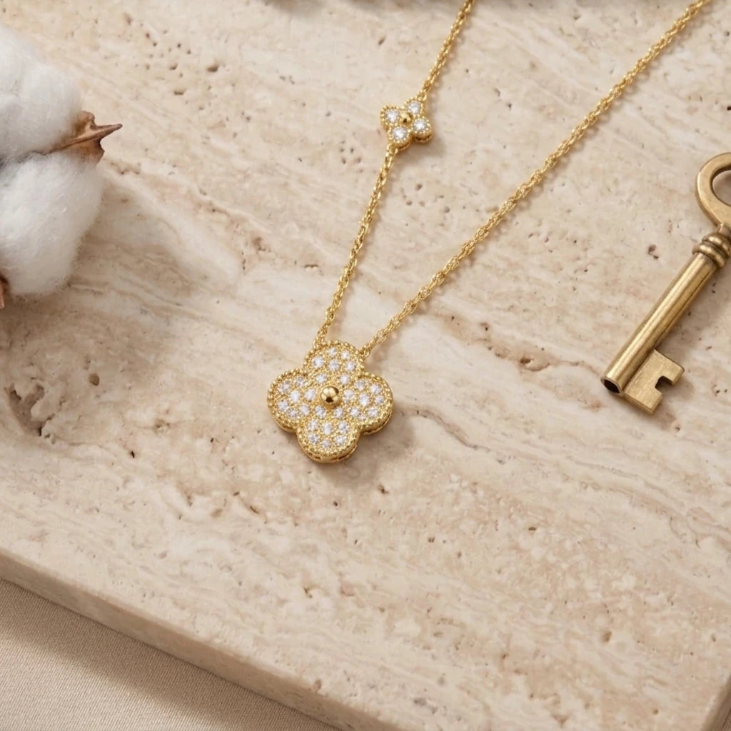 Vue de dessus en gros plan d'un délicat collier doré reposant sur une plaque de marbre travertin beige texturé. Le collier est doté d'un pendentif principal en forme de trèfle à quatre feuilles entièrement serti de diamants pavés étincelants, avec une petite perle centrale dorée. Une station plus petite, en forme de fleur à quatre pétales, est visible plus haut sur la chaîne. La scène est décorée d'une cosse de coton blanche et douce sur la gauche et d'une clé d'époque en laiton patiné sur la droite. 