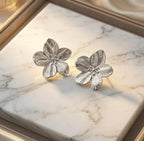 Paire de boucles d'oreilles fleurs argentées Léonie Majestueuse posées sur une plaque de marbre blanc veiné. La lumière rasante souligne les détails ciselés des pétales et la finition brillante du métal.