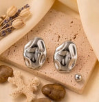 Paire de boucles d'oreilles argentées Léna - Aquatique posée sur un support en pierre beige, soulignant leur forme fluide et leur éclat solaire.