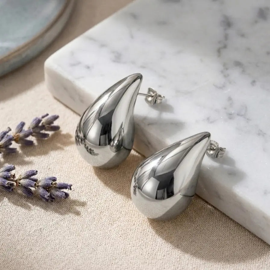 Paire de boucles d'oreilles Inès Irrésistible en finition argentée, posée sur un bloc de marbre blanc à côté de brins de lavande séchée. Rendu luxe et réaliste.