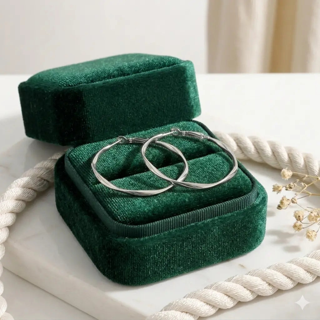 Paire de boucles d'oreilles créoles torsadées argentée, modèle Élise Élégante, présentées dans un écrin de velours vert émeraude. Le métal argenté scintille sur le support en marbre blanc.
