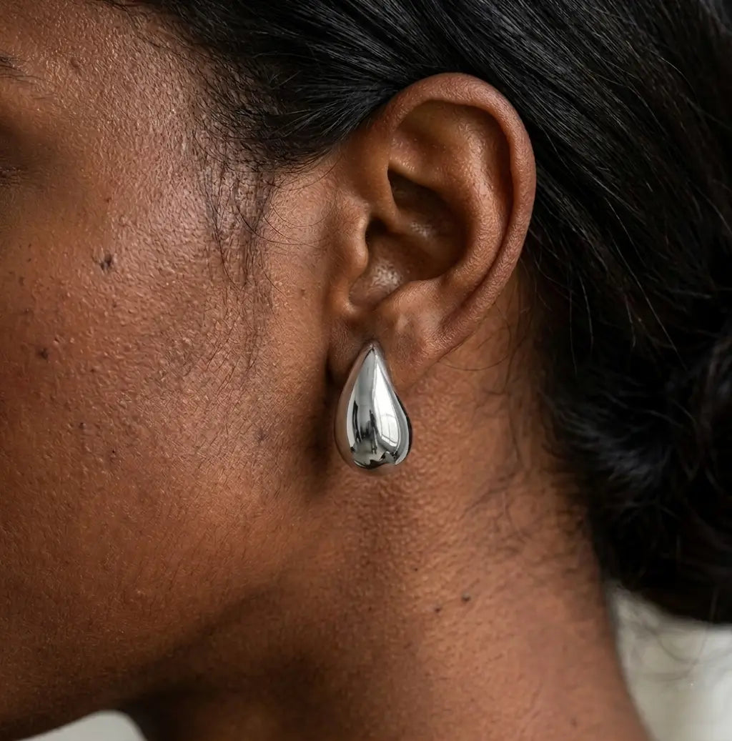 Gros plan réaliste d'une boucle d'oreille goutte argenté poli, modèle Inès Irrésistible, portée à l'oreille. Le métal reflète la lumière sur une peau texturée.