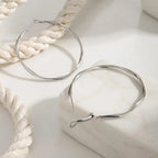 Gros plan sur une paire de boucles d'oreilles créoles torsadées argentées, modèle Élise Élégante. Une boucle repose sur un socle en marbre blanc, montrant le fermoir tige et la structure fine et ondulée. L'autre boucle est à l'arrière-plan, près d'un cordon de coton blanc tressé. Photographie lumineuse avec un éclairage doux.