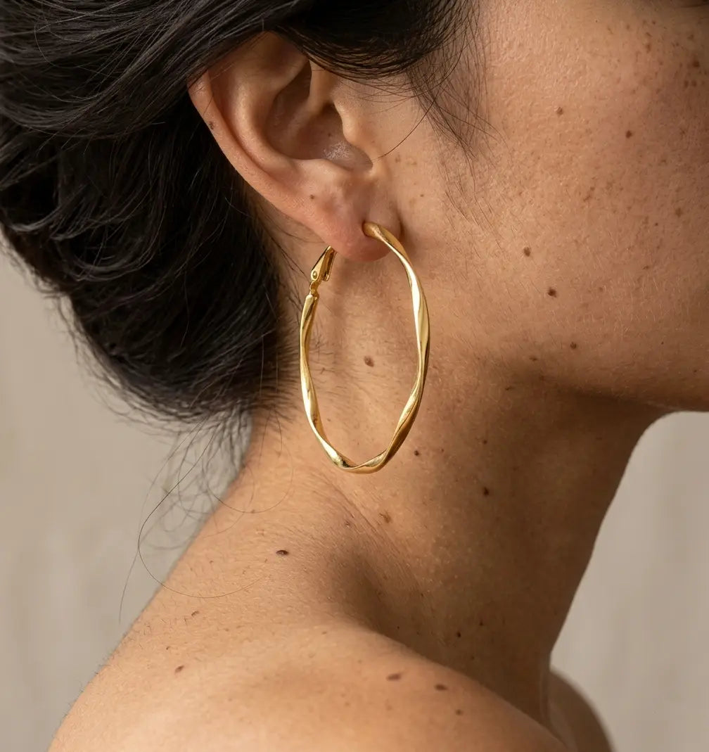 Gros plan réaliste d'une boucle d'oreille en acier inoxydable or Élise Élégante portée par une mannequin. On distingue les détails de la peau et le mouvement torsadé du bijou argenté qui apporte un éclat sophistiqué.