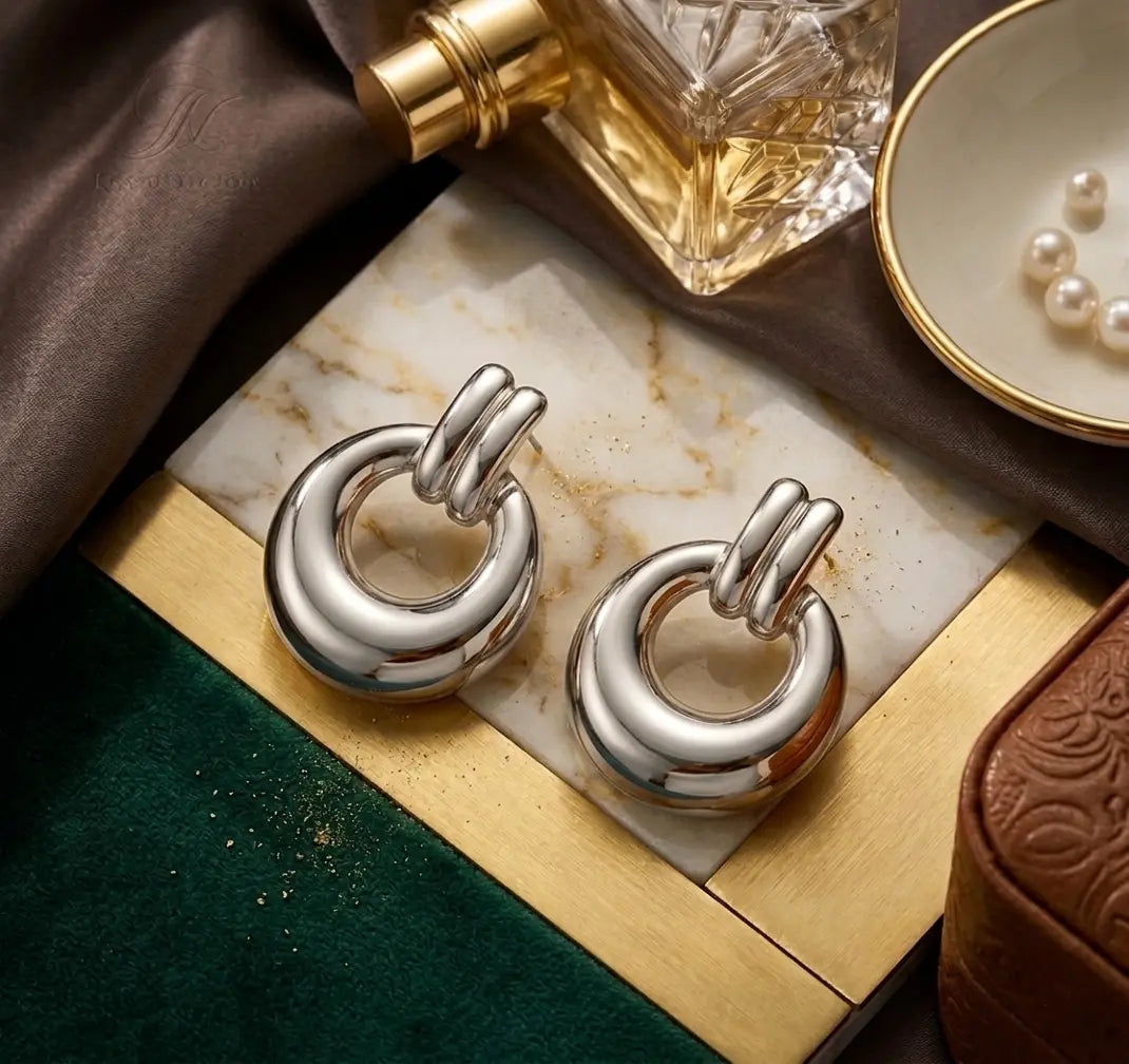 Paire de boucles d'oreilles massives Alba argentées posée sur un plateau en marbre et laiton. La scène chic inclut un parfum de luxe et un plat avec des perles.