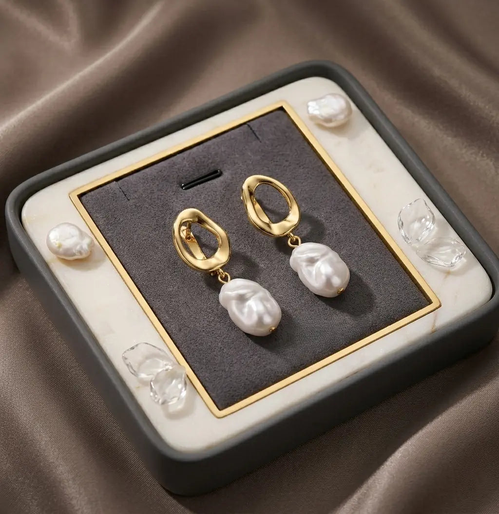 Vue du dessus des boucles d'oreilles Clarisse - Nacrée dorées dans leur écrin minimaliste en velours gris avec des détails dorés. Le packaging haut de gamme prêt à offrir.