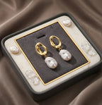 Vue du dessus des boucles d'oreilles Clarisse - Nacrée dorées dans leur écrin minimaliste en velours gris avec des détails dorés. Le packaging haut de gamme prêt à offrir.