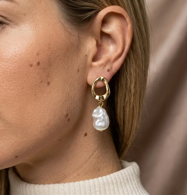 Plan rapproché d'un mannequin de profil portant la boucle d'oreille Clarisse - Nacrée dorée. L'anneau torsadé or et la perle baroque d'eau douce illuminent le visage sur un fond de soie taupe.