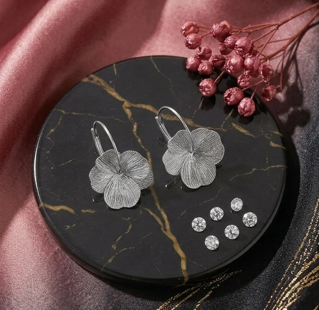 Vue à plat de deux boucles d'oreilles Eléonore en acier inoxydable argenté brossé reposant sur un disque de marbre noir avec des fleurs séchées et de petits zircons.
