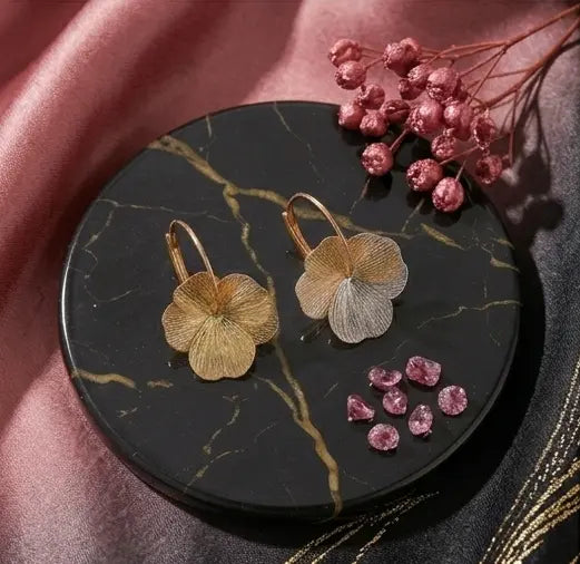 Vue à plat de deux boucles d'oreilles Eléonore en acier inoxydable doré brossé reposant sur un disque de marbre noir avec des fleurs séchées et de petits zircons.