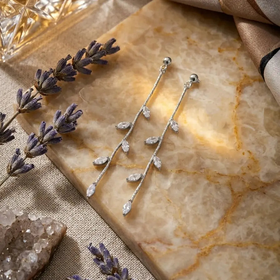 Paire de boucles d'oreilles pendantes Héloïse-Divine argentées, délicatement posée sur une plaque de marbre veiné. La scène est décorée de brins de lavande séchée, de cristaux bruts et d'un tissu de soie, dans une lumière chaude et douce.