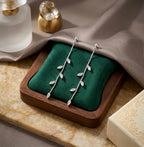 Gros plan de la paire de boucles d'oreilles pendantes Héloïse-Divine en acier inoxydable dorées, reposant sur un coussin en velours vert émeraude à l'intérieur d'un écrin en bois. La composition chic inclut un flacon de parfum givré et de la soie grise.