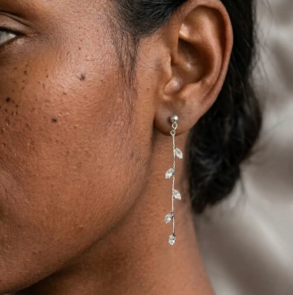 Gros plan sur l'oreille d'un modèle portant la boucle d'oreille pendante Héloïse-Divine argentées. La boucle est fine, avec une chaîne dorée ornée de cristaux en forme de feuilles qui scintillent près du visage.