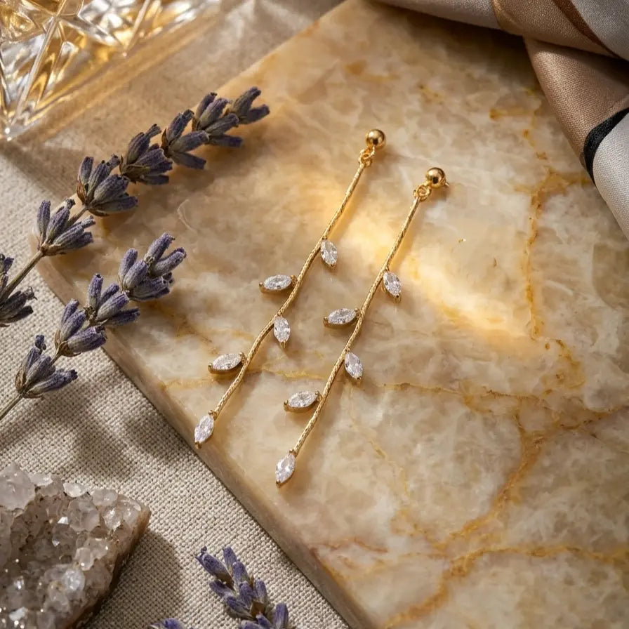 Paire de boucles d'oreilles pendantes Héloïse-Divine en or, délicatement posée sur une plaque de marbre veiné. La scène est décorée de brins de lavande séchée, de cristaux bruts et d'un tissu de soie, dans une lumière chaude et douce. 