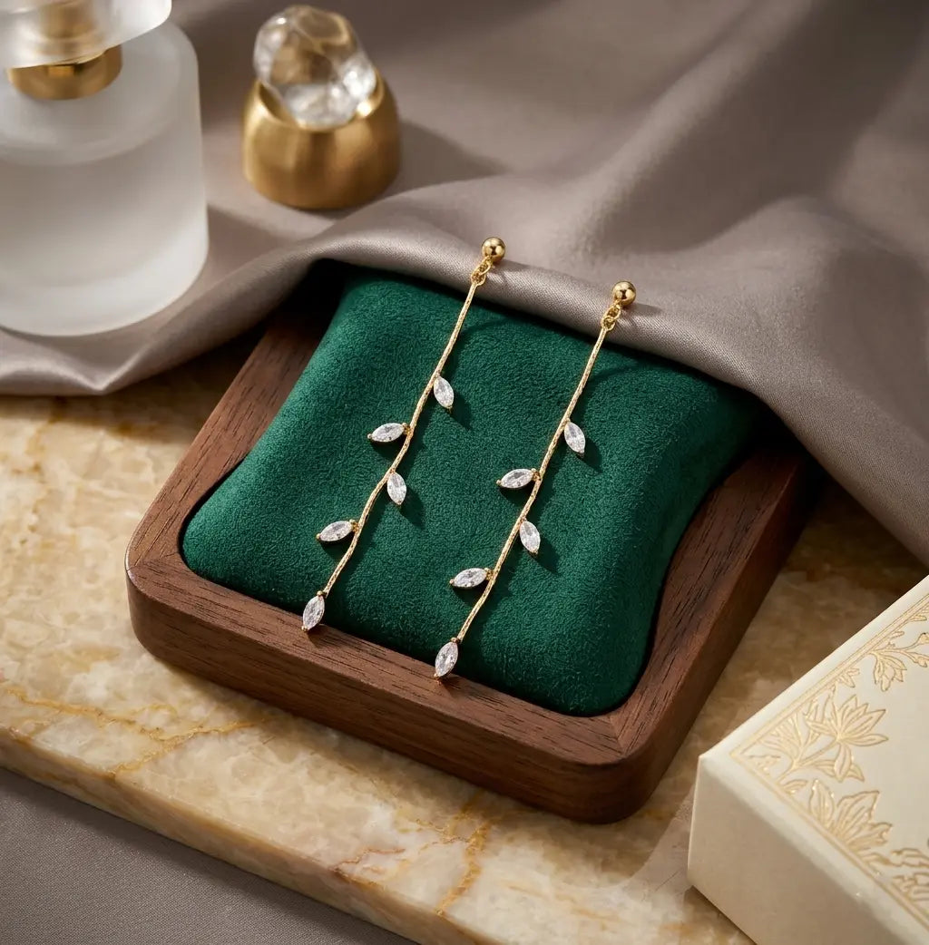 Gros plan de la paire de boucles d'oreilles pendantes Héloïse-Divine en acier inoxydable dorées, reposant sur un coussin en velours vert émeraude à l'intérieur d'un écrin en bois. La composition chic inclut un flacon de parfum givré et de la soie grise.