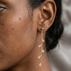 Gros plan sur l'oreille d'un modèle portant la boucle d'oreille pendante Héloïse-Divine en acier inoxydable or. La boucle est fine, avec une chaîne dorée ornée de cristaux en forme de feuilles qui scintillent près du visage.