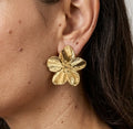 Gros plan sur le profil d'une femme portant une boucle d'oreille "Léonie Majestueuse" doré. Le bijou, en forme de fleur épanouie aux pétales finement texturés et striés, apporte une touche d'élégance naturelle.