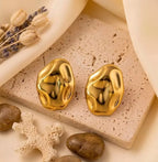 Paire de boucles d'oreilles dorées Léna - Aquatique posée sur un support en pierre beige, soulignant leur forme fluide et leur éclat solaire.