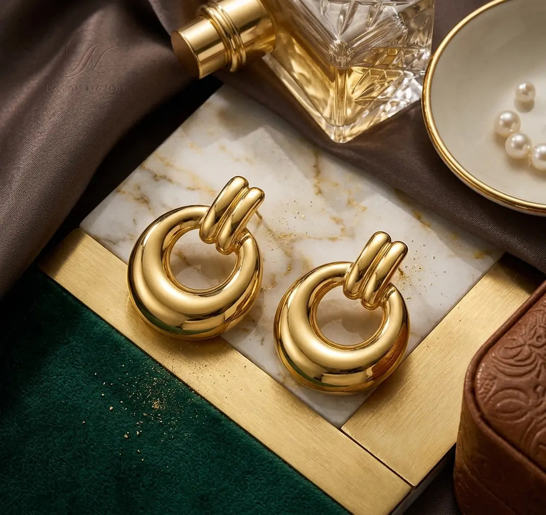 Paire de boucles d'oreilles massives Alba dorées posée sur un plateau en marbre et laiton. La scène chic inclut un parfum de luxe et un plat avec des perles.