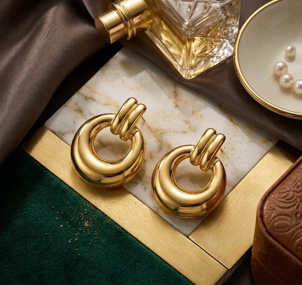 Paire de boucles d'oreilles massives Alba dorées posée sur un plateau en marbre et laiton. La scène chic inclut un parfum de luxe et un plat avec des perles.