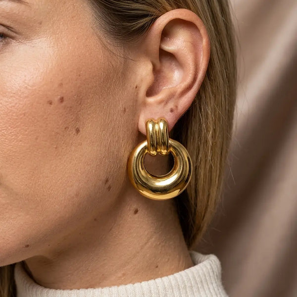 Gros plan sur le lobe d'oreille d'un modèle portant la boucle d'oreille dorée massive Alba. Le bijou sculptural est net, tandis que le visage et l'arrière-plan en soie sont floutés.