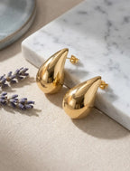Paire de boucles d'oreilles Inès Irrésistible en finition dorée, posée sur un bloc de marbre blanc à côté de brins de lavande séchée. Rendu luxe et réaliste.