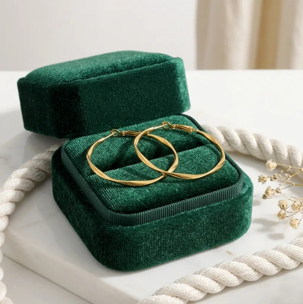 Paire de boucles d'oreilles créoles torsadées en acier inoxydable or, modèle Élise Élégante, présentées dans un écrin de velours vert émeraude. Le métal argenté scintille sur le support en marbre blanc.