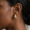 Gros plan réaliste d'une boucle d'oreille goutte en acier inoxydable poli, modèle Inès Irrésistible, portée à l'oreille. Le métal reflète la lumière sur une peau texturée.