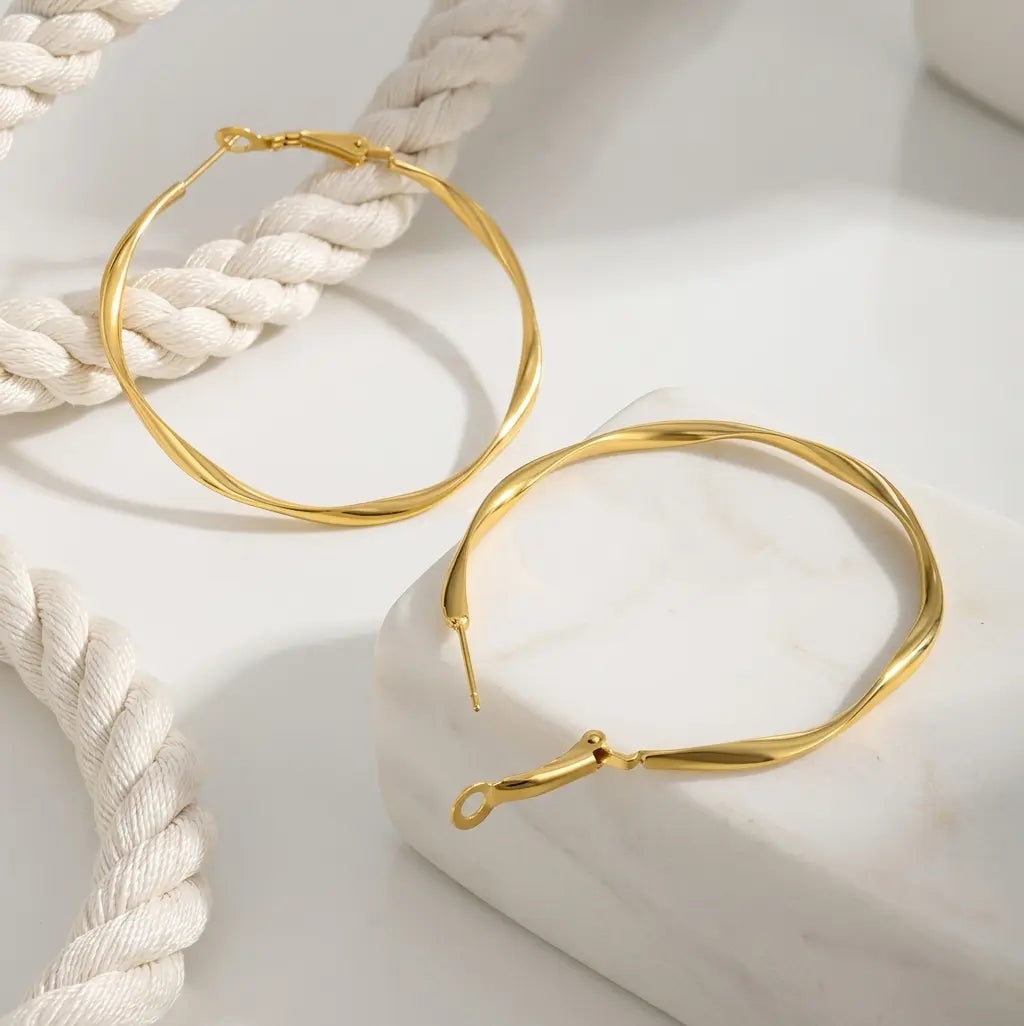 Gros plan sur une paire de boucles d'oreilles créoles torsadées en acier inoxydable or, modèle Élise Élégante. Une boucle repose sur un socle en marbre blanc, montrant le fermoir tige et la structure fine et ondulée. L'autre boucle est à l'arrière-plan, près d'un cordon de coton blanc tressé. Photographie lumineuse avec un éclairage doux.