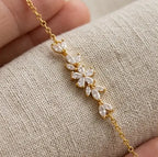 Gros plan détaillé sur le motif central floral en oxydes de zirconium taille marquise du bracelet Chloé de Maison Dosana, suspendu au-dessus d'un tissu en lin texturé. L'acier inoxydable doré et les zircons brillent sous la lumière.