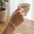 gros plan sur le poignet d'une femme portant le bracelet Chloé en acier inoxydable doré et oxydes de zirconium de Maison Dosana, tenant une tasse de thé vintage. Le bracelet délicat scintille au poignet.