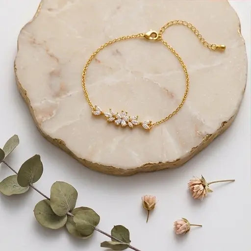 Vue d'ensemble à plat du bracelet Chloé - Étincelante de Maison Dosana reposant sur une plaque de marbre rose avec des fleurs séchées. Le bracelet en acier inoxydable doré présente un motif floral central en zircons brillants.