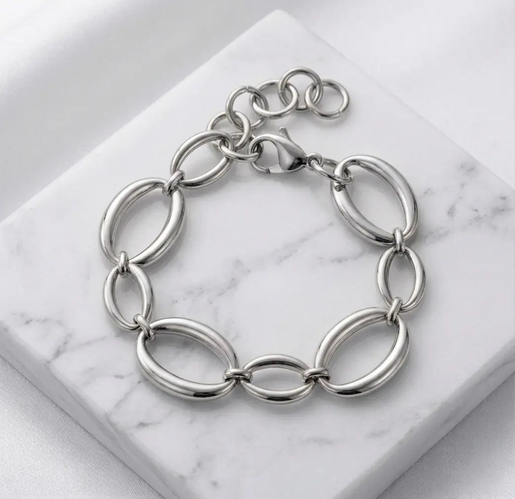 Photographie à plat (flat lay) du bracelet argenté Victoire Vibrante. Il est posé sur un bloc de marbre blanc veiné, sur un fond de satin blanc. Les maillons ovales et le fermoir mousqueton sont bien visibles.