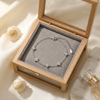 Présentation cadeau du bracelet argenté Margaux Fine dans son coffret de joaillerie en bois naturel. Le couvercle en verre laisse entrevoir le bijou reposant sur un coussinet protecteur, entouré d'accessoires de luxe suggérant un produit haut de gamme.