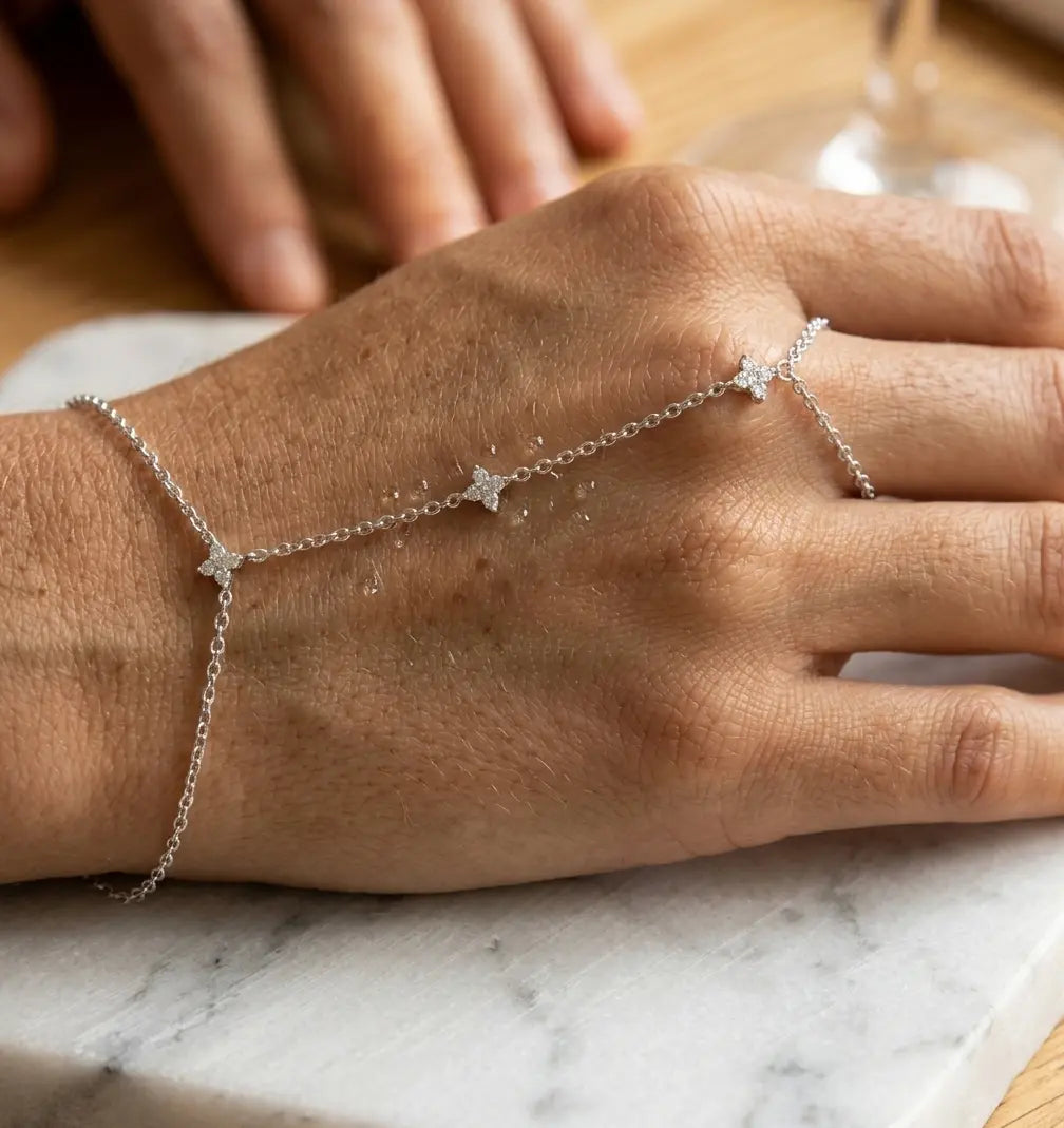 Gros plan macro du bracelet de main Lola Libre en argent sterling 925, sur marbre de Carrare. Des gouttes d'eau sont délicatement posées sur la chaîne fleurie et scintillante, évoquant la pureté et l'éclat naturel. Le bijou semble flotter sur la main.