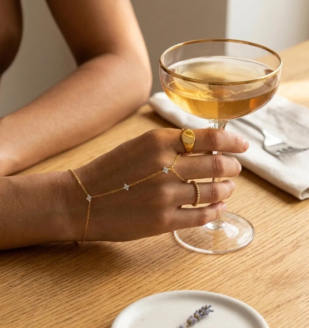 La mannequin porte le bracelet de main Lola Libre en plaqué or, habillant sa main d'un éclat délicat. Elle tient un verre de cocktail sur une table en bois brossé. Cette parure de main fleurie apporte une touche d'élégance à votre style.
