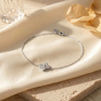 Gros plan du bracelet Sia - Cristalline argenté posé sur un tissu en satin crème et une pierre de travertin. Le pendentif trèfle à quatre feuilles est serti de cristaux étincelants qui captent la lumière chaude du soleil.