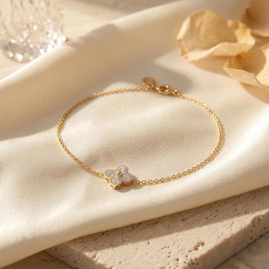 Gros plan du bracelet Sia - Cristalline doré posé sur un tissu en satin crème et une pierre de travertin. Le pendentif trèfle à quatre feuilles est serti de cristaux étincelants qui captent la lumière chaude du soleil.