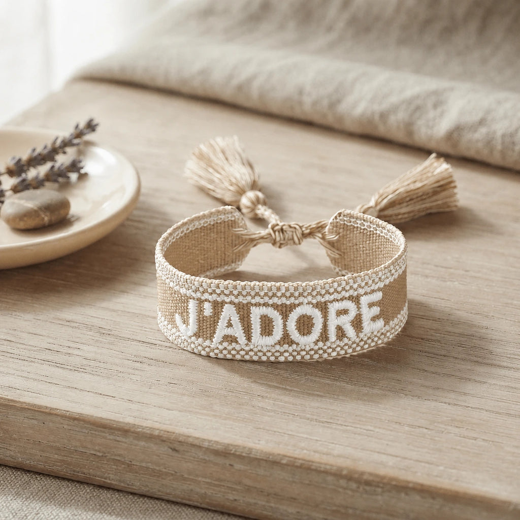 Bracelet tissé de couleur beige avec l'inscription 'J'ADORE', posé sur une planche en bois clair, accompagné d'une petite coupelle contenant des brins de fleurs séchées de lavande et un galet.