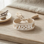 Bracelet tissé de couleur beige avec l'inscription 'J'ADORE', posé sur une planche en bois clair, accompagné d'une petite coupelle contenant des brins de fleurs séchées de lavande et un galet.