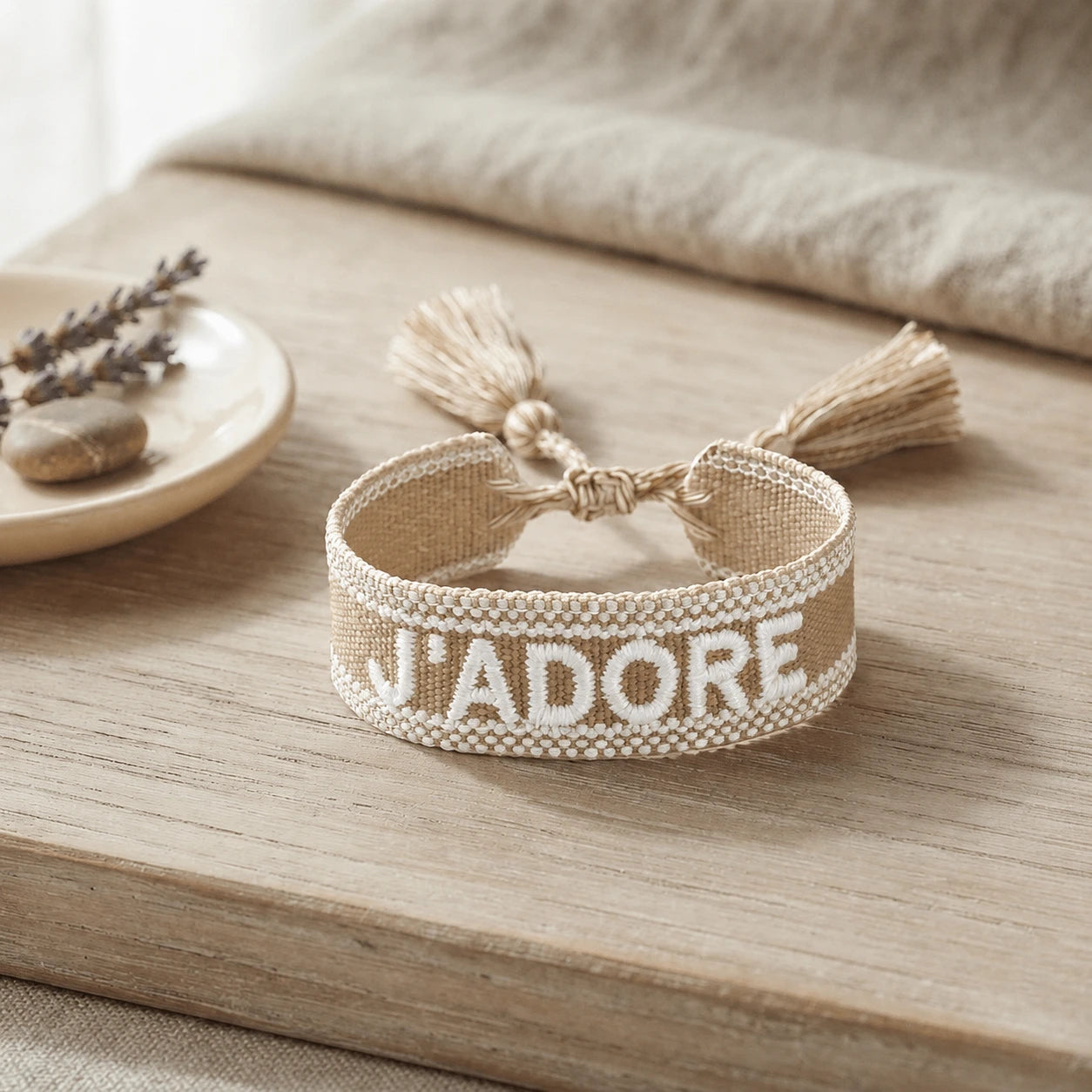 Bracelet tissé de couleur beige avec l'inscription 'J'ADORE', posé sur une planche en bois clair, accompagné d'une petite coupelle contenant des brins de fleurs séchées de lavande et un galet.