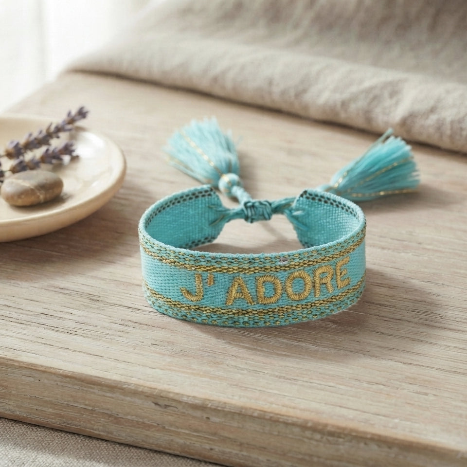 Bracelet tissé de couleur bleu avec l'inscription 'J'ADORE', posé sur une planche en bois clair, accompagné d'une petite coupelle contenant des brins de fleurs séchées de lavande et un galet.