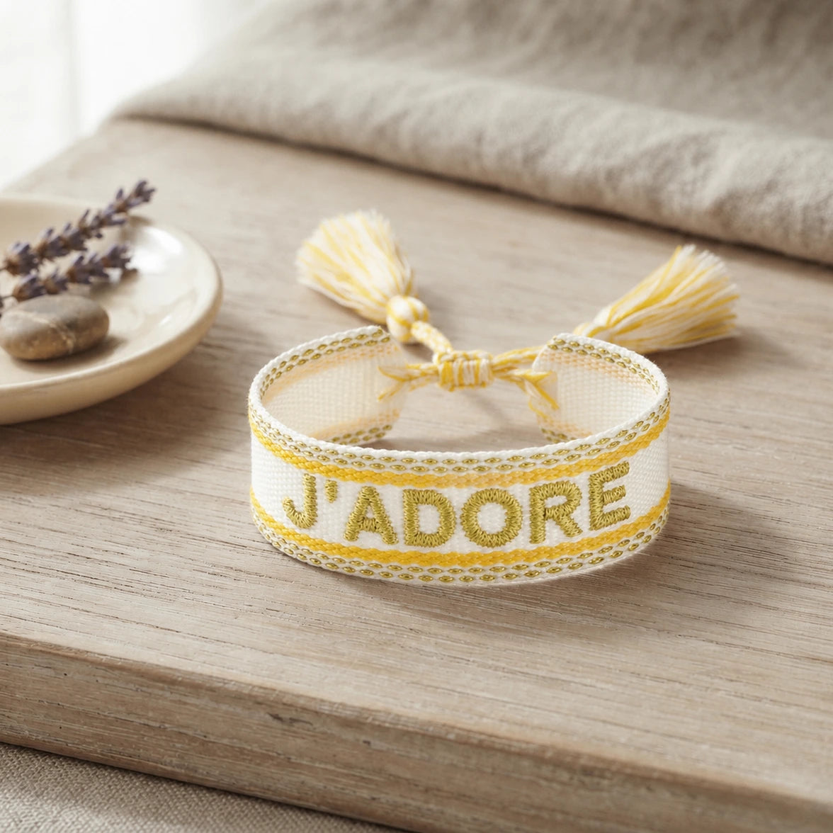 Bracelet tissé de couleur jaune avec l'inscription 'J'ADORE', posé sur une planche en bois clair, accompagné d'une petite coupelle contenant des brins de fleurs séchées de lavande et un galet.