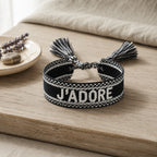 Bracelet tissé de couleur noir avec l'inscription 'J'ADORE', posé sur une planche en bois clair, accompagné d'une petite coupelle contenant des brins de fleurs séchées de lavande et un galet.