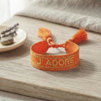 Bracelet tissé de couleur orange avec l'inscription 'J'ADORE', posé sur une planche en bois clair, accompagné d'une petite coupelle contenant des brins de fleurs séchées de lavande et un galet.