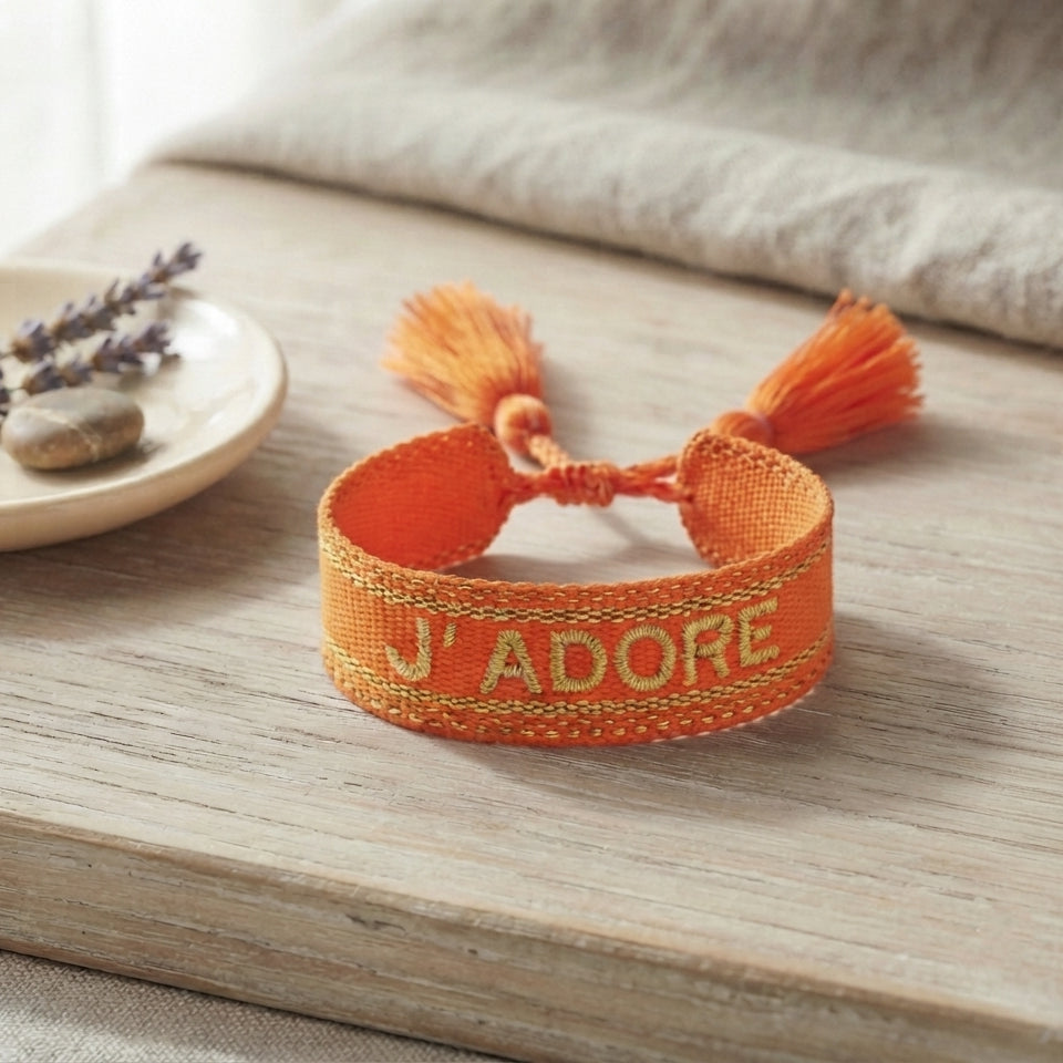 Bracelet tissé de couleur orange avec l'inscription 'J'ADORE', posé sur une planche en bois clair, accompagné d'une petite coupelle contenant des brins de fleurs séchées de lavande et un galet.